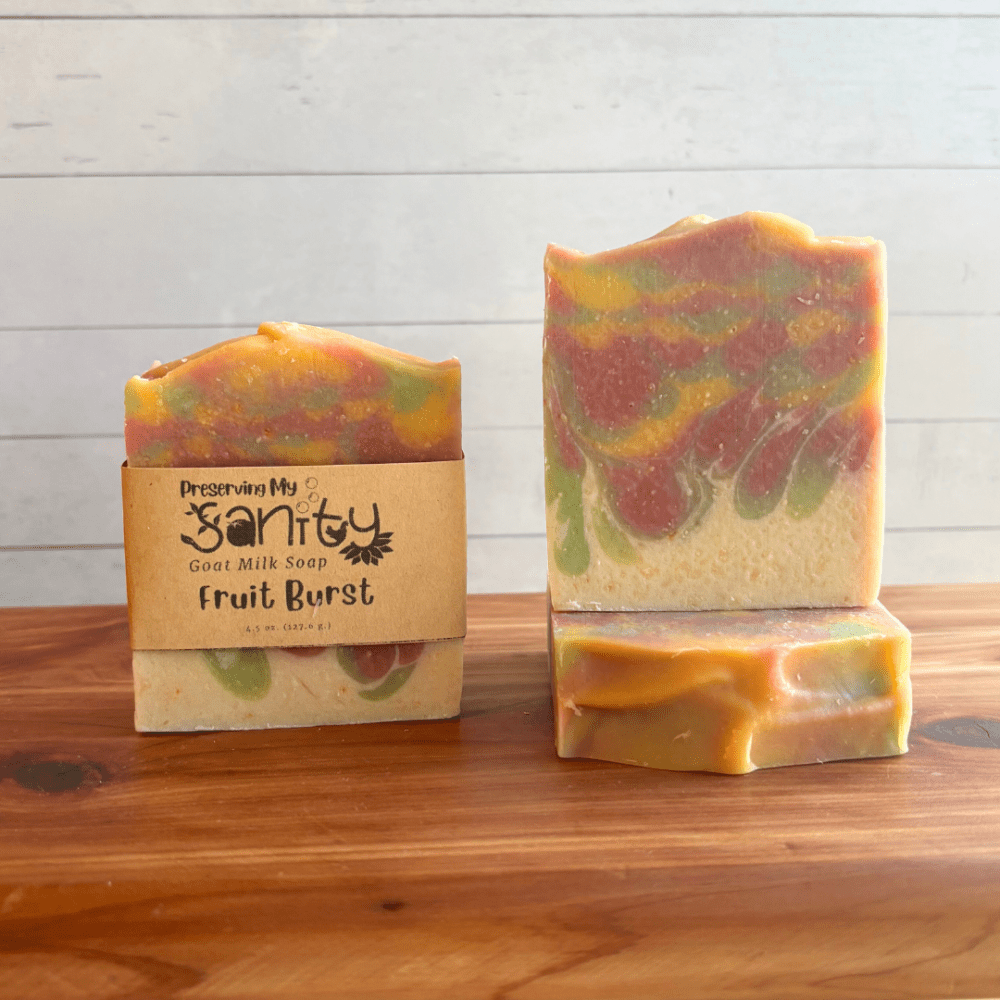 Alternate view display photo showcasing three bars of Fruit Burst goat milk soap sitting on a cedar board with a white shiplap background. This colorful soap has white on the bottom and is topped with a unique swirl of green, red, and yellow. It smells like fruity gum!