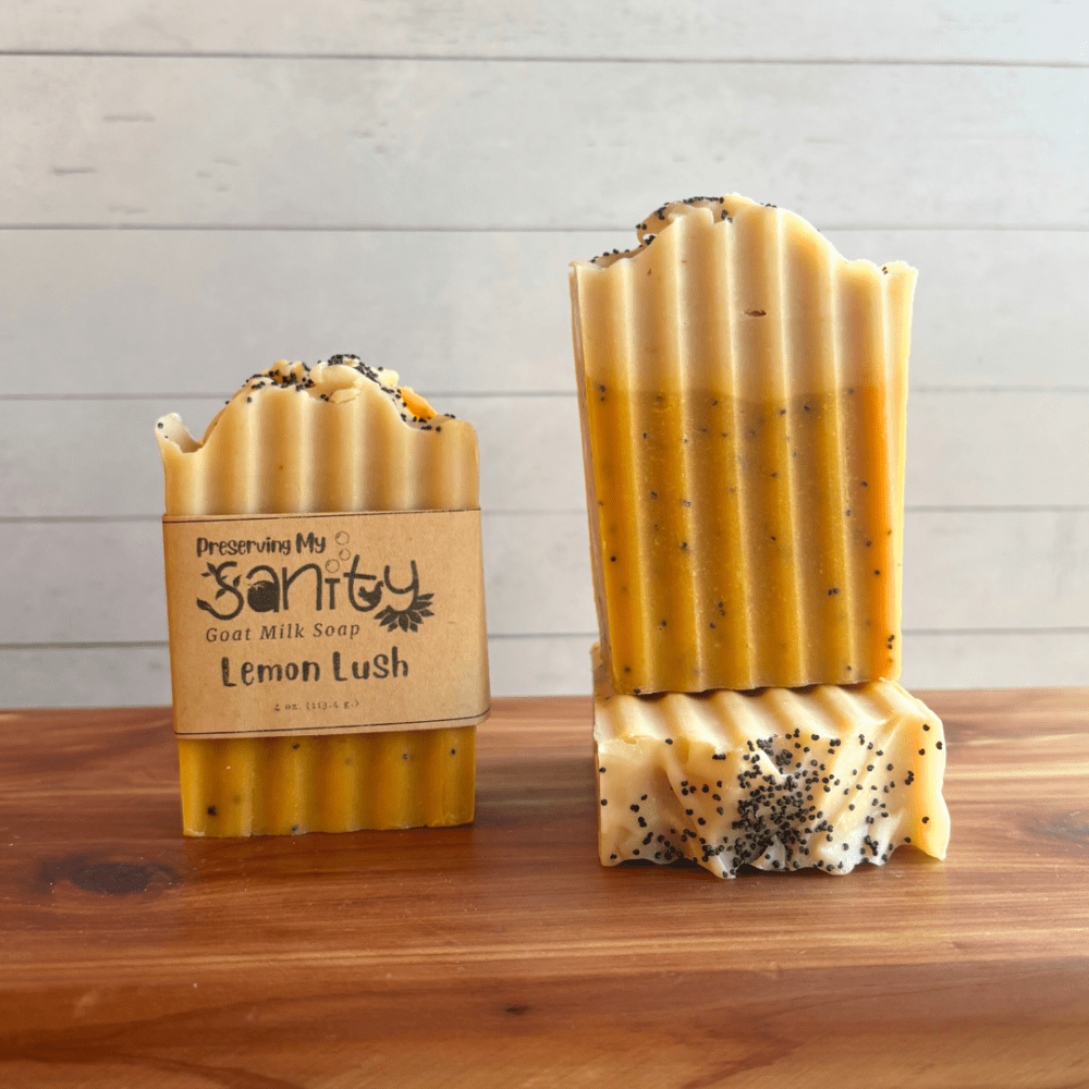 Alternate view display photo showcasing three bars of Lemon Lush goat milk soap sitting on a cedar board with a white shiplap background. The soap was poured in two layers, with a medium yellow as the main base color that fills 3/4 of the soap, topped with white - like frosting in a lemon lush dessert. There are poppyseeds throughout the soap for exfoliation and on top, and these bars were cut with a wavy ridged cutter.