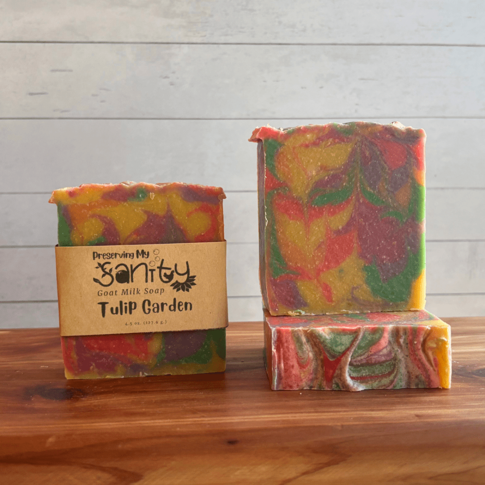 Alternate view display photo showcasing three bars of Tulip Garden goat milk soap, in colorful pretty swirls of pink, purple, green, yellow, and orange. Soaps are sitting on a cedar bath board with a white shiplap background.