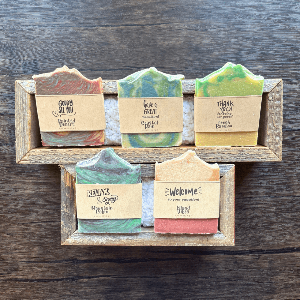 Flatlay photo showcasing five half-size bars of soap to be used as vacation rental guest welcome soaps. They are displayed in two reclaimed wood boxes with a white crocheted washcloth behind them. Scents include Painted Desert, Coastal Rain, Fresh Bamboo, Mountain Cabin, and Island Vibes. They are sitting on a dark wood background.