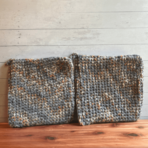 Two crocheted potholders in a gray and beige variegated pattern situated on a cedar bath board with a white panel background.
