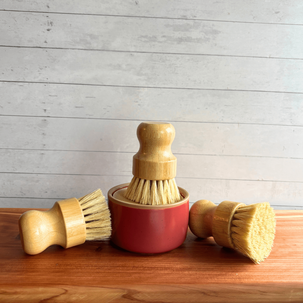 A photo showing one red ceramic ramekin with a dish soap bar pictured with three bamboo handle dish brushes. Listing is for the purchase of one dish brush, but the photo shows multiple so potential buyers can see all angles.