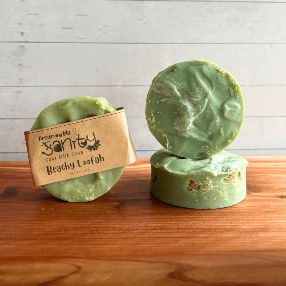 Three bars of beachy loofah soap sitting on a cedar bath board. One bar has a brand label, the other two are shown without labels and are situated to show the top and side of the round bars of soap, which are colored a pretty green.