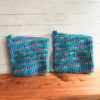 Set of two blue crocheted potholders sitting on a display board with white paneling in the background. The blue is an aqua color and it's mixed with a variegated multi color yarn to make this colorful set.