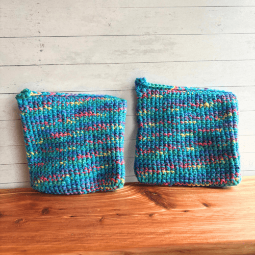 Set of two blue crocheted potholders sitting on a display board with white paneling in the background. The blue is an aqua color and it's mixed with a variegated multi color yarn to make this colorful set.