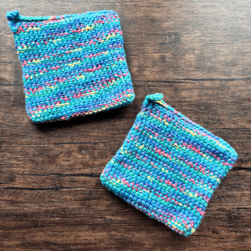 Set of two variegated blue crocheted potholders arranged separately on a dark piece of wood as the backdrop.