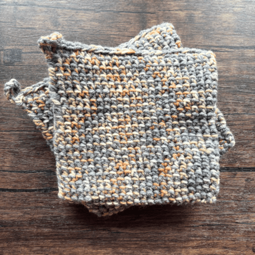 Set of two variegated gray and beige crocheted potholders sitting on top of each other with a dark piece of wood as the backdrop.