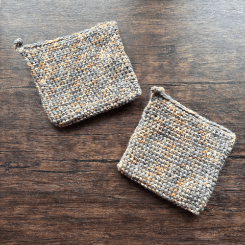 Set of two variegated gray and beige crocheted potholders arranged separately on a dark piece of wood as the backdrop.
