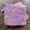 Set of two variegated purple crocheted potholders sitting on top of each other with a dark piece of wood as the backdrop.