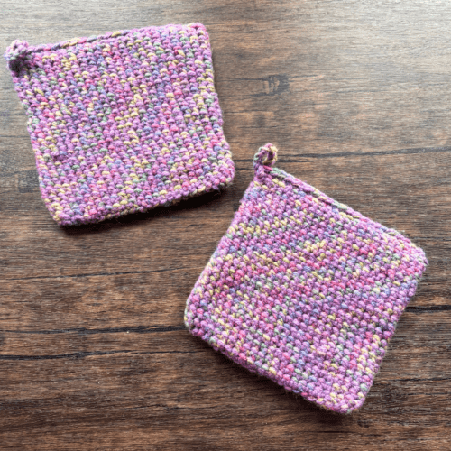 Set of two variegated purple crocheted potholders arranged separately on a dark piece of wood as the backdrop.