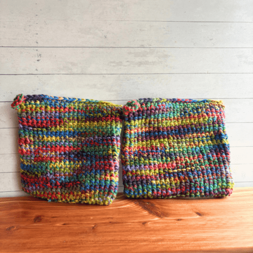 Set of two rainbow crocheted potholders sitting on a display board with white paneling in the background.