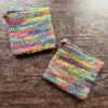 Set of two variegated rainbow crocheted potholders arranged separately on a dark piece of wood as the backdrop.