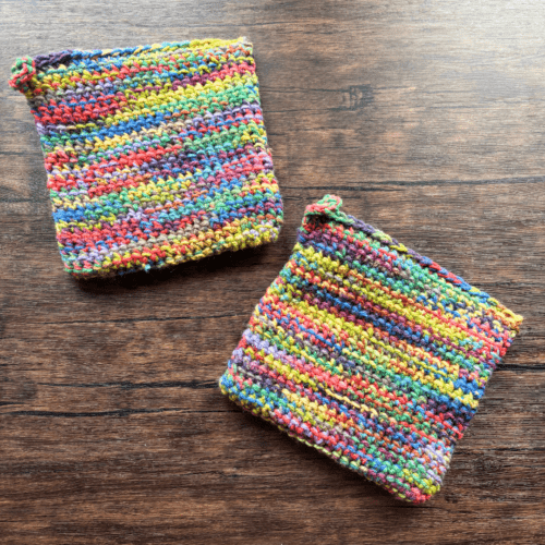 Set of two variegated rainbow crocheted potholders arranged separately on a dark piece of wood as the backdrop.