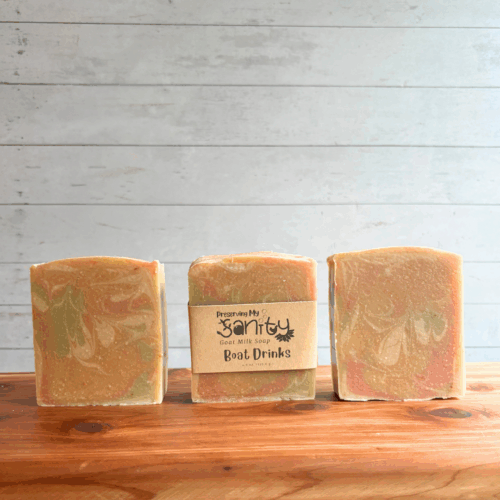 Three bars of Boat Drinks goat milk soap sitting in a row on a cedar board with white panel background. The soap itself has pretty swirls of white, green, pink, and orange in real muted shades. One bar of soap has a label on it, the other two don't so you can see the whispy swirls.