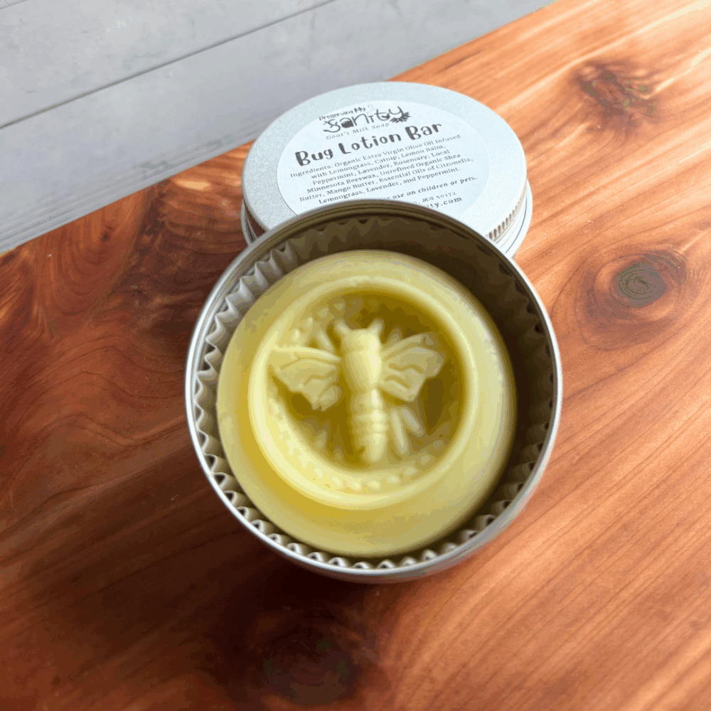 Display photo showing a bug lotion bar, infused with herbs like lemongrass, catnip, peppermint, and lavender and featuring essential oils of citronella, lemongrass, and peppermint. The lotion bar has an indent of a bee in it from the silicon mold it's poured into and is shown in a reusable aluminum tin.
