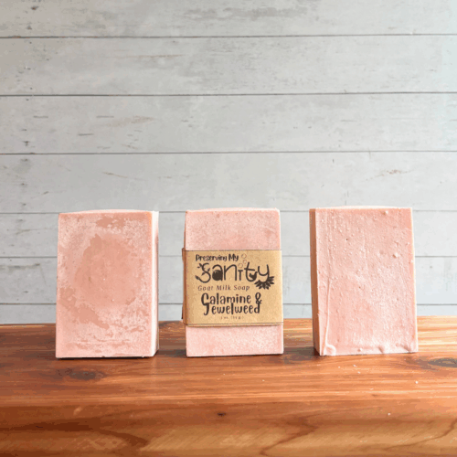 Three bars of Calamine & Jewelweed soap, shown with the middle bar labeled and the other two unlabeled to show the soaps pretty pink color and plain design. Sitting on a cedar board with white panel background.