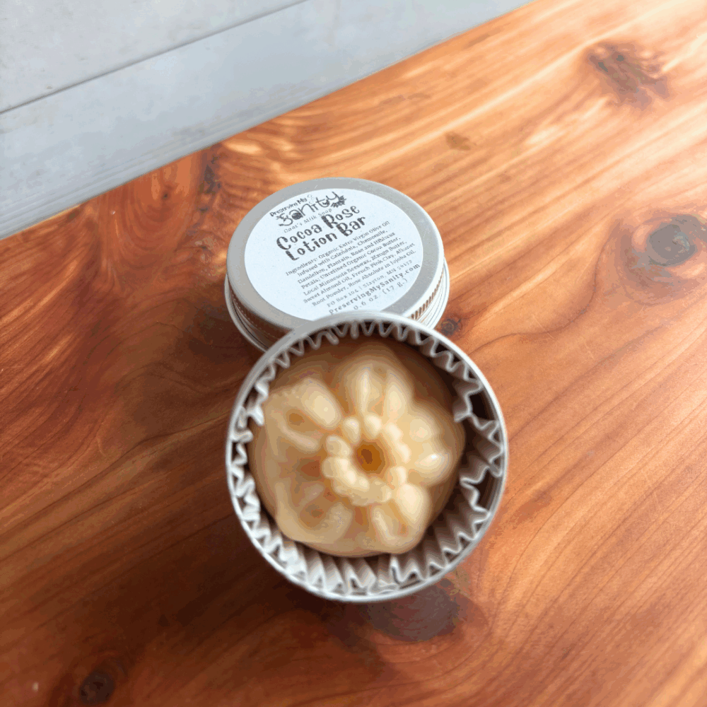Display photo of a Cocoa Rose lotion bar in a reusable aluminum tin. The solid lotion bar is a pretty flower shape and is a light pink color, with the labeled lid in the background. Sitting on a cedar board with a white paneled background.