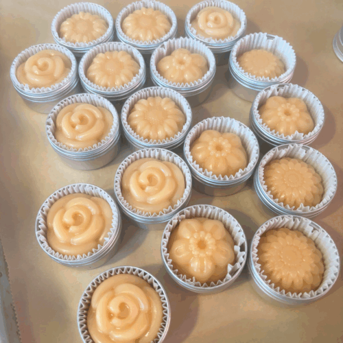 Process photo showing a tray of freshly made Cocoa Rose Lotion Bars, just taken out of the silicon flower mold and placed into reusable aluminum tins. The bars are various flower shapes and a pretty pink color.