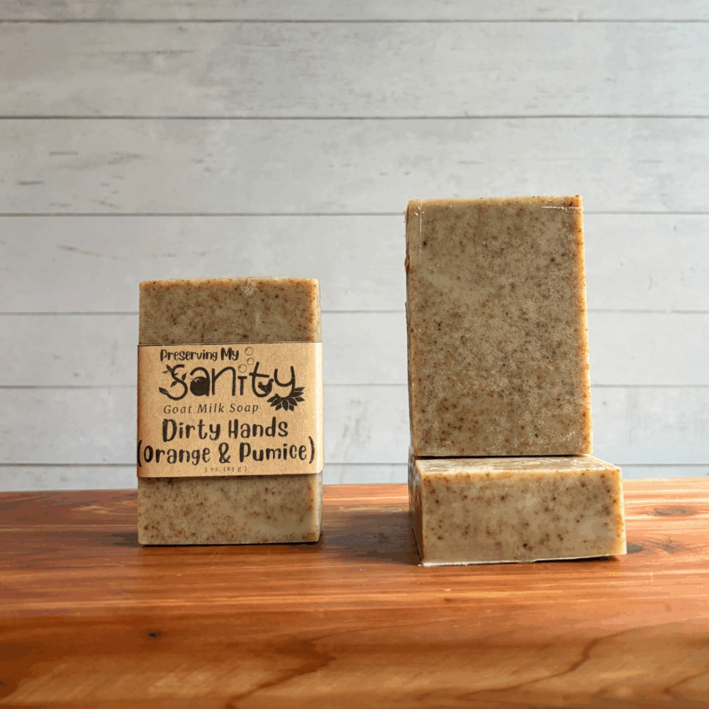Three bars of Fresh Lilac goat milk soap. The middle bar has a label, while the other two are unlabeled to show the color and plain design of this exfoliating orange pumice soap. All are sitting on a cedar board with a white paneled background.