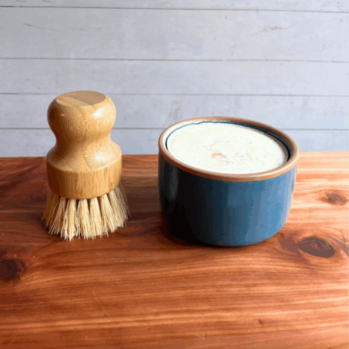Dish soap set showing a bar of cold process basil and citrus dish soap in a blue ceramic ramekin with a bamboo dish brush. Both are sitting on a cedar board with a white panel background.