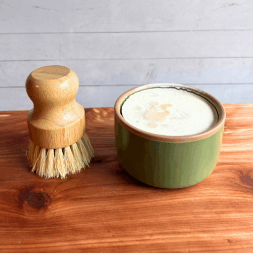 Dish soap set showing a bar of cold process basil and citrus dish soap in a green ceramic ramekin with a bamboo dish brush. Both are sitting on a cedar board with a white panel background.