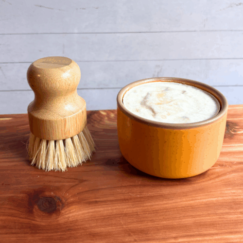Dish soap set showing a bar of cold process basil and citrus dish soap in a yellow ceramic ramekin with a bamboo dish brush. Both are sitting on a cedar board with a white panel background.