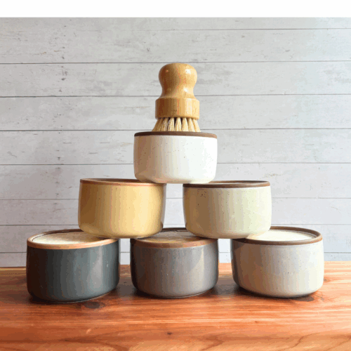 Display photo showcasing six ceramic ramekins of dish soap bars in neutral colors, with a bamboo dish brush sitting on top of the pyramid style display.