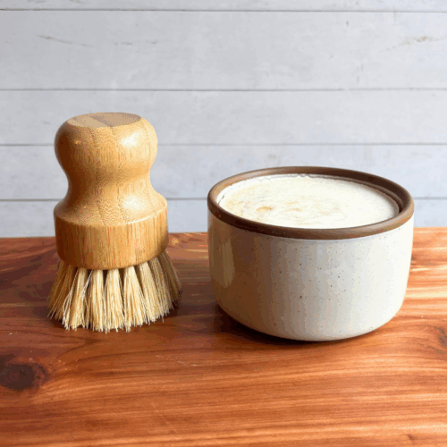 Dish soap set showing a bar of cold process basil and citrus dish soap in a ceramic ramekin with a bamboo dish brush. Both are sitting on a cedar board with a white panel background.