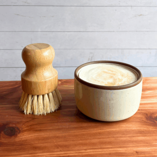 Dish soap set showing a bar of cold process basil and citrus dish soap in a ceramic ramekin with a bamboo dish brush. Both are sitting on a cedar board with a white panel background.