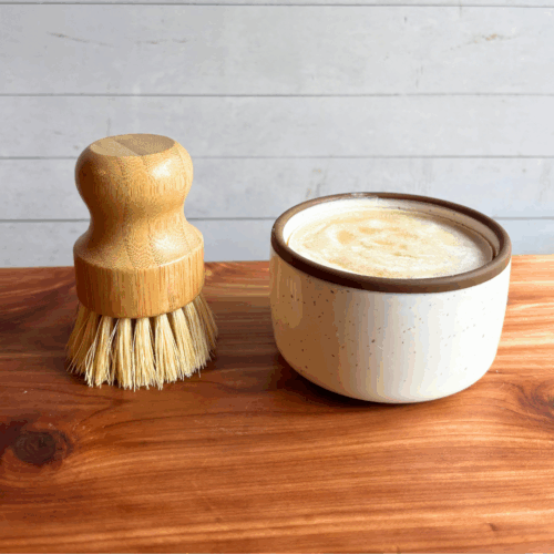 Dish soap set showing a bar of cold process basil and citrus dish soap in a ceramic ramekin with a bamboo dish brush. Both are sitting on a cedar board with a white panel background.