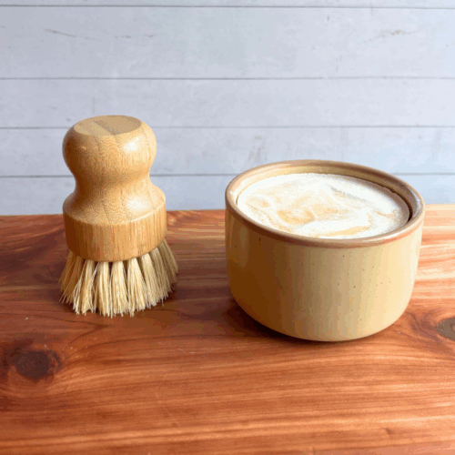 Dish soap set showing a bar of cold process basil and citrus dish soap in a tan ceramic ramekin with a bamboo dish brush. Both are sitting on a cedar board with a white panel background.
