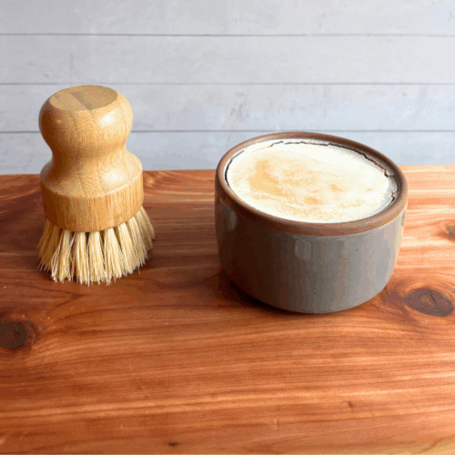 Dish soap set showing a bar of cold process basil and citrus dish soap in a taupe ceramic ramekin with a bamboo dish brush. Both are sitting on a cedar board with a white panel background.
