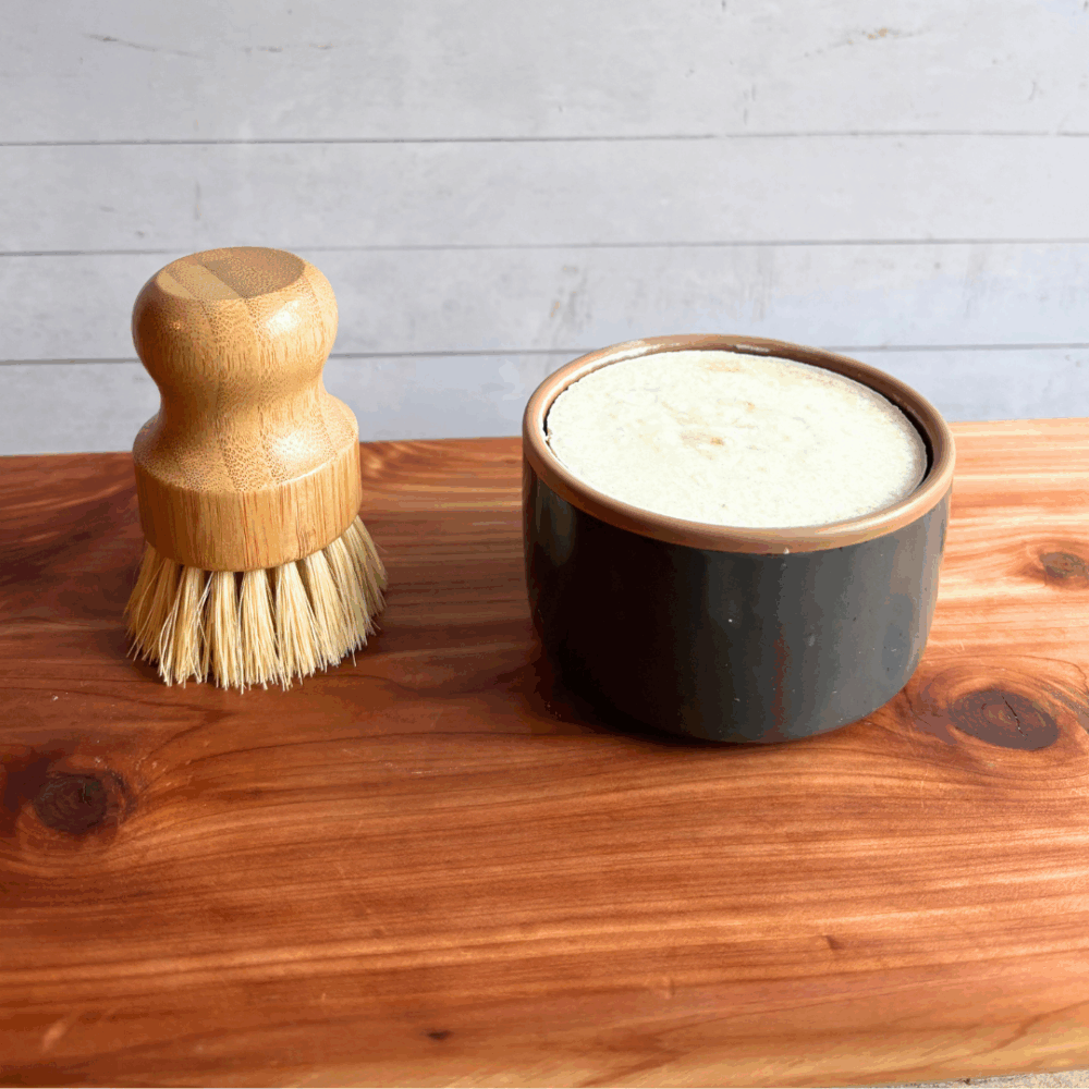 Dish soap set showing a bar of cold process basil and citrus dish soap in a gray ceramic ramekin with a bamboo dish brush. Both are sitting on a cedar board with a white panel background.