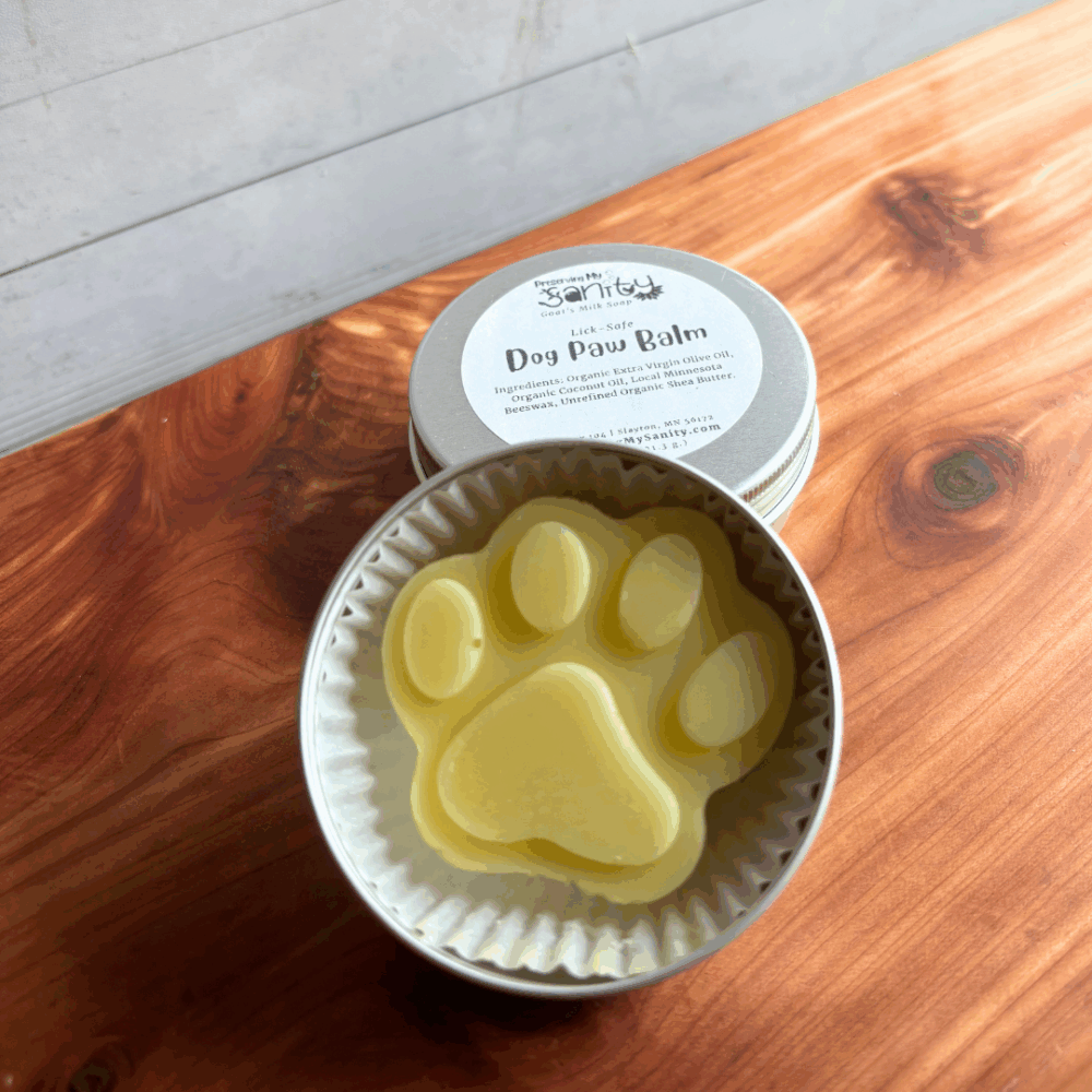 Display photo showing a paw-shaped dog paw balm in a reusable aluminum tin with a label in the background. Made in small batches in my Minnesota kitchen, these are perfect for dog paws and noses. Sitting on a cedar board with white panel background.