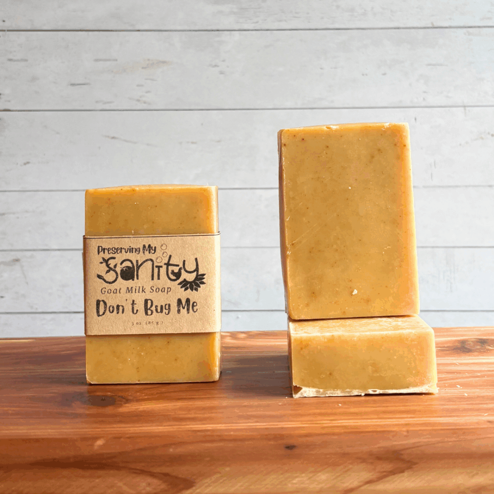 Three bars of Don't Bug Me goat milk soap. The left bar has a label, while the other two are unlabeled to show the yellow color and plain design of this soap. All are sitting on a cedar board with a white paneled background.