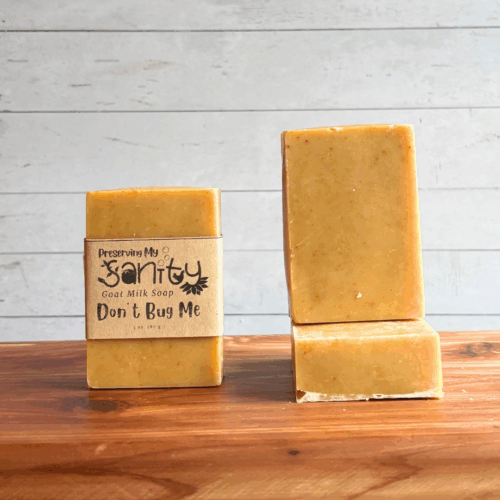 Three bars of Don't Bug Me goat milk soap. The left bar has a label, while the other two are unlabeled to show the yellow color and plain design of this soap. All are sitting on a cedar board with a white paneled background.