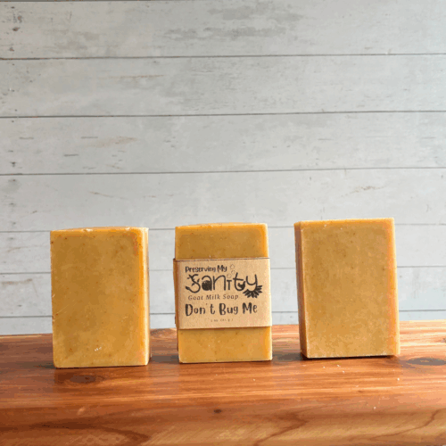 Three bars of Don't Bug Me goat milk soap. The middle bar has a label, while the other two are unlabeled to show the yellow color and plain design of this soap. All are sitting on a cedar board with a white paneled background.