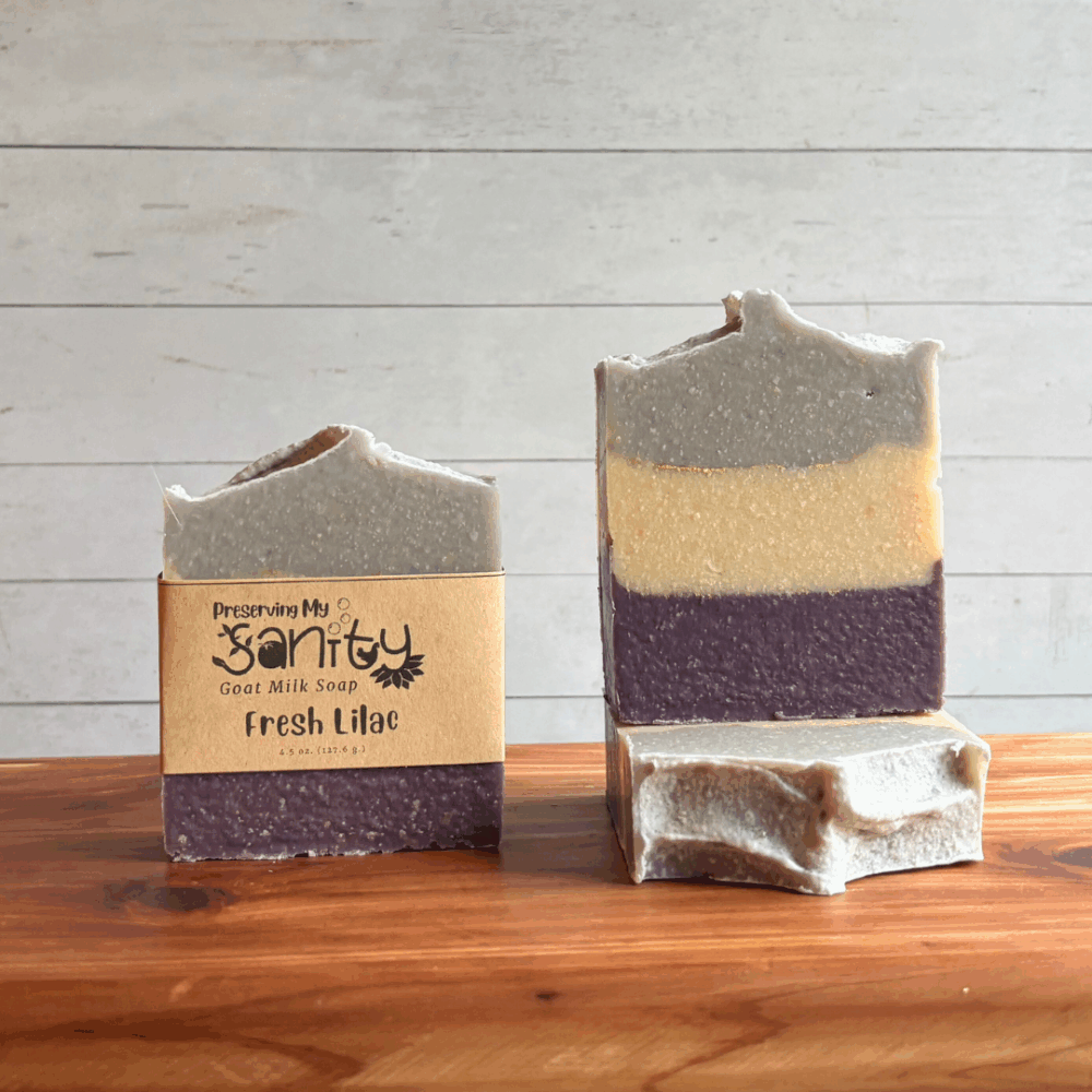 Three bars of Fresh Lilac goat milk soap. The left bar has a label, while the other two are unlabeled to show the top and striped design of purple, white, and lavender with gold mica stripes between layers. All are sitting on a cedar board with a white paneled background.
