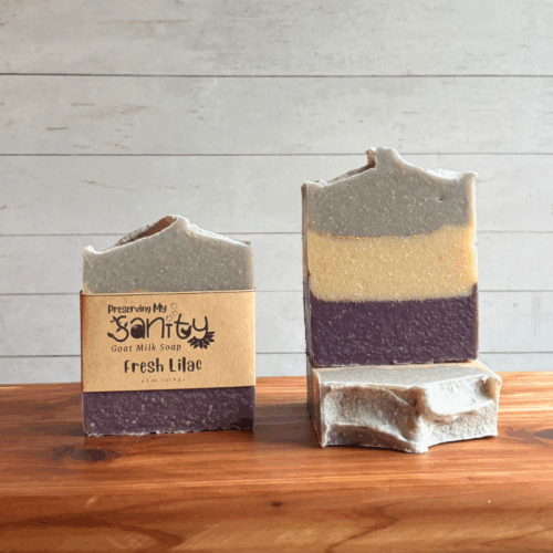 Three bars of Fresh Lilac goat milk soap. The left bar has a label, while the other two are unlabeled to show the top and striped design of purple, white, and lavender with gold mica stripes between layers. All are sitting on a cedar board with a white paneled background.