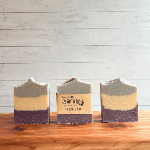 Three bars of Fresh Lilac goat milk soap. The middle bar has a label, while the other two are unlabeled to show the striped design of purple, white, and lavender with gold mica stripes between layers. All are sitting on a cedar board with a white paneled background.