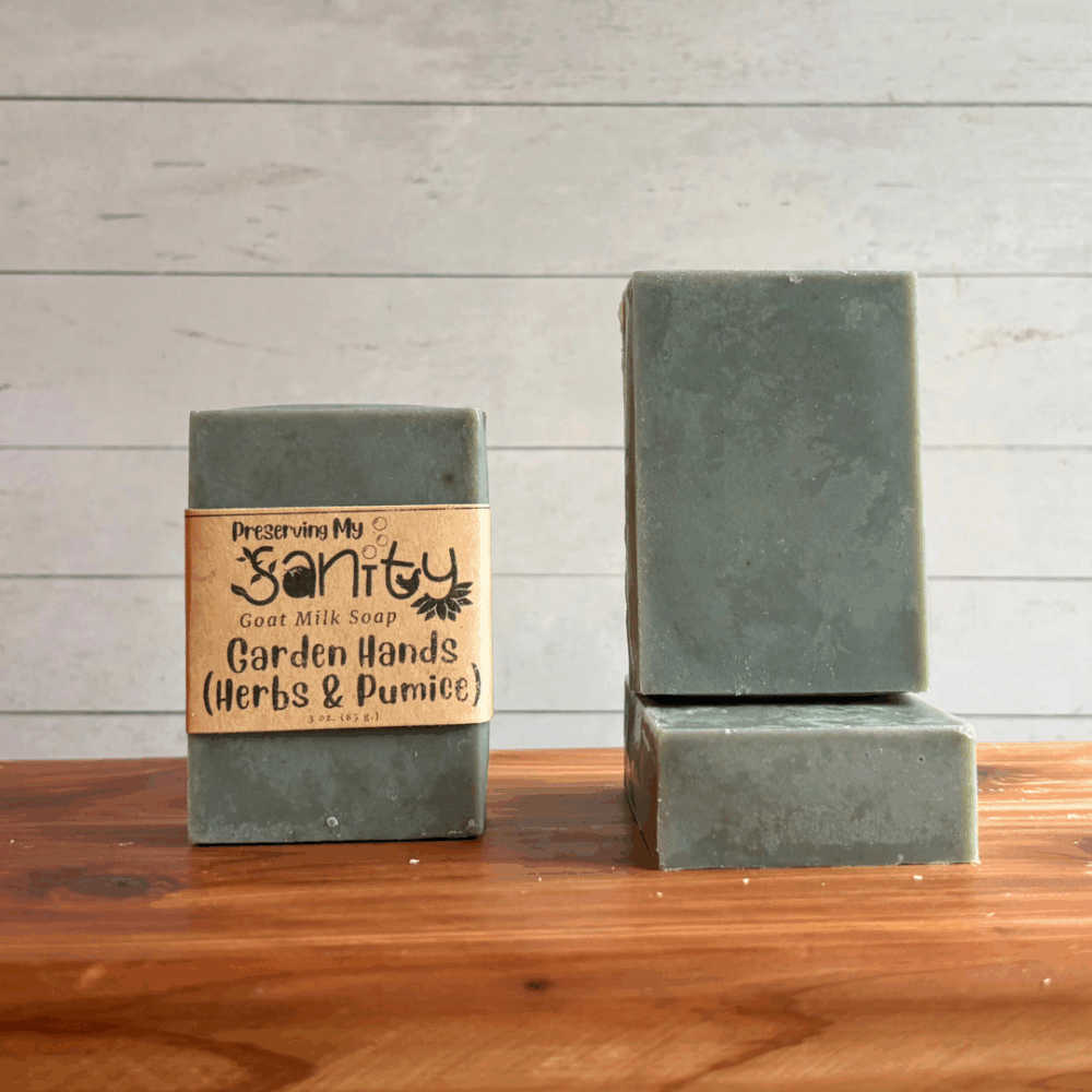 Three bars of Garden Hands goat milk soap. The left bar has a label, while the other two are unlabeled to show the gray/green color of this exfoliating goat milk soap. All are sitting on a cedar board with a white paneled background.