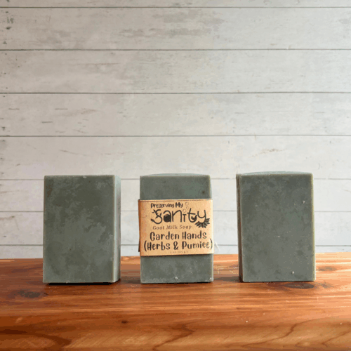 Three bars of Garden Hands goat milk soap. The middle bar has a label, while the other two are unlabeled to show the gray/green color of this exfoliating goat milk soap. All are sitting on a cedar board with a white paneled background.