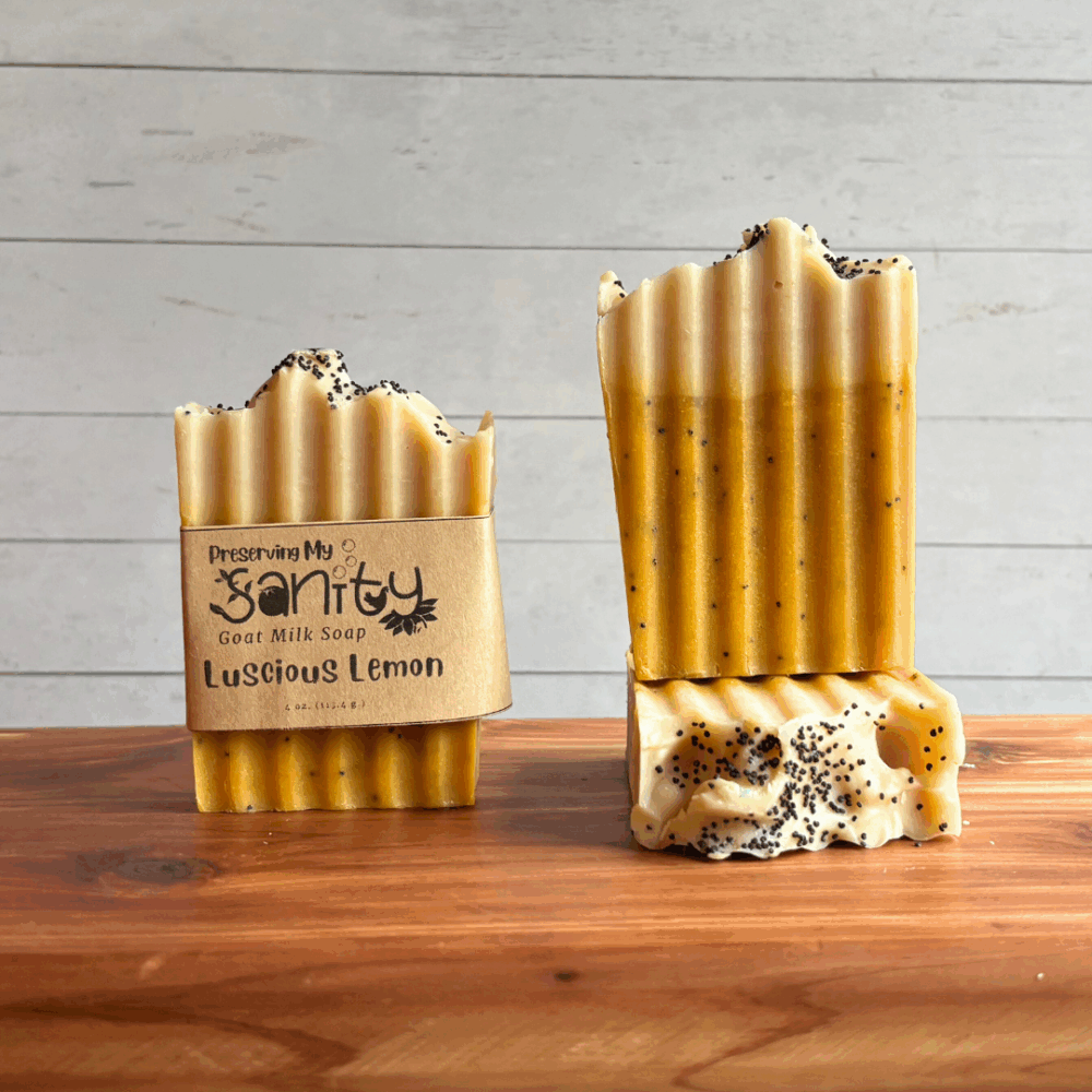 Three bars of Luscious Lemon poppyseed soap, pictured on a cedar board with a white paneled background. The left bar has a label, while the other two are unlabeled to show the yellow soap design with a white top.