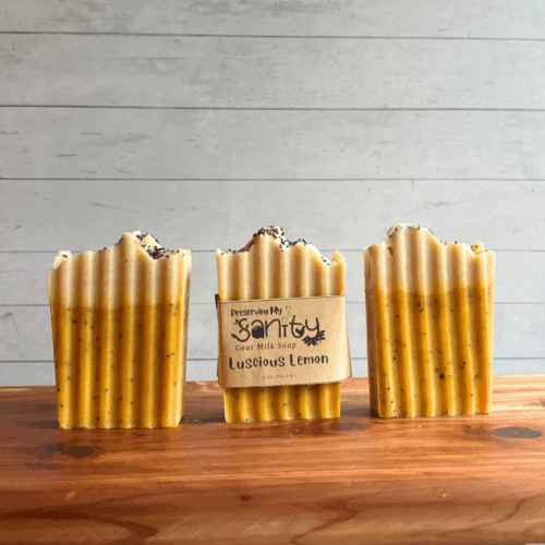 Three bars of Luscious Lemon poppyseed soap, pictured on a cedar board with a white paneled background. The middle bar has a label, while the other two are unlabeled to show the yellow soap design with a white top.