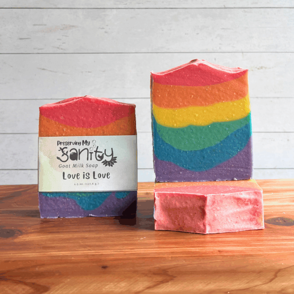 Three bars of rainbow Love is Love goat milk soap, displayed on a cedar board with a white panel background to show the label on one bar, top of one bar, and front face of the third.