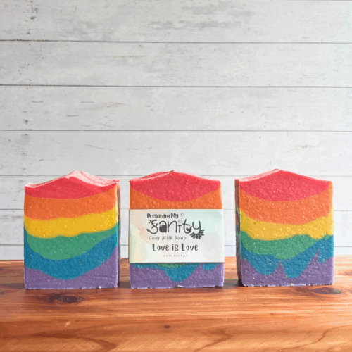 Three colorful rainbow bars of Love is Love goat milk soap, featuring the six colors of the Pride flag in a striped pattern perfect for celebrating Pride Month. The middle bar has a label, while the other two are unlabeled to show soap design. All are sitting on a cedar board with a white paneled background.