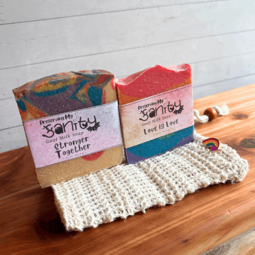 Alternate view display photo showing two half-size bars of Pride-themed soap in a soap saver bag with a Pride lapel pin. These items make up the Pride Sampler, a great gift to show your support of Pride Month. Pictured on a cedar board with a white paneled background.