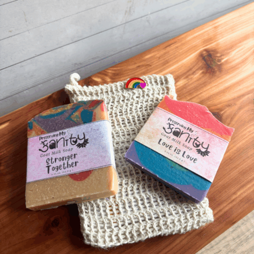 Alternate view display photo showing two half-size bars of Pride-themed soap in a soap saver bag with a Pride lapel pin. These items make up the Pride Sampler, a great gift to show your support of Pride Month. Pictured on a cedar board with a white paneled background.
