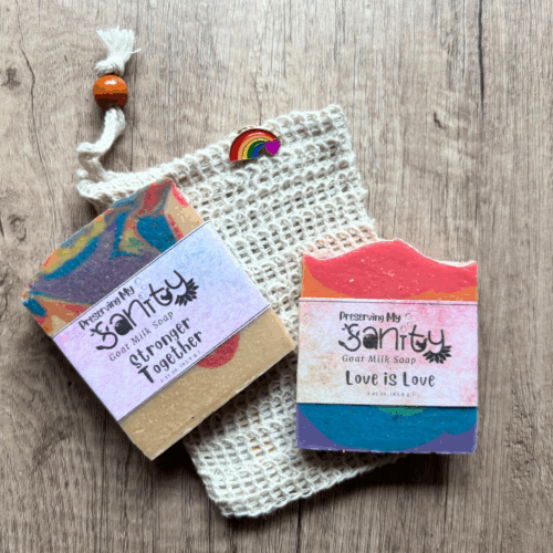 Flatlay photo showing two half-size bars of Pride-themed soap in a soap saver bag with a Pride lapel pin. These items make up the Pride Sampler, a great gift to show your support of Pride Month. Pictured on a wood grain background board.