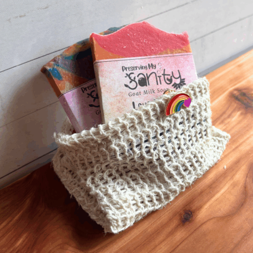 Display photo showing two half-size bars of Pride-themed soap in a soap saver bag with a Pride lapel pin. These items make up the Pride Sampler, a great gift to show your support of Pride Month. Pictured on a cedar board with a white paneled background.
